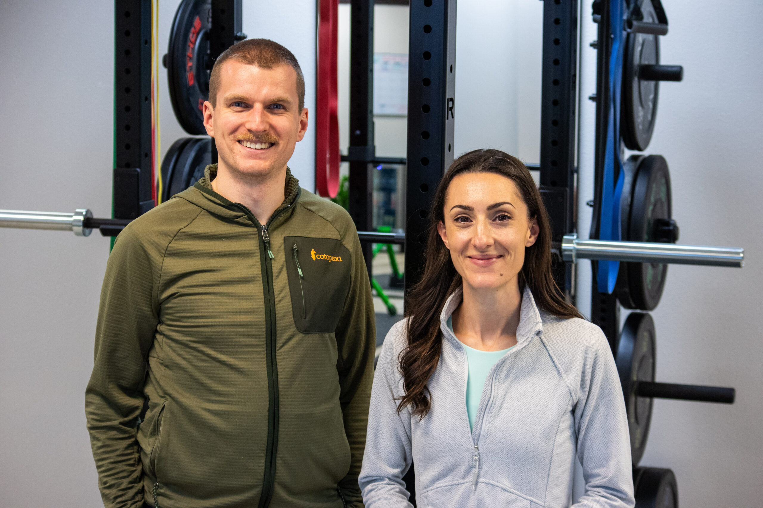 Physical therapists Ben Hoskyn and Maria Leibler standing side by side and smiling in front of a squat rack.