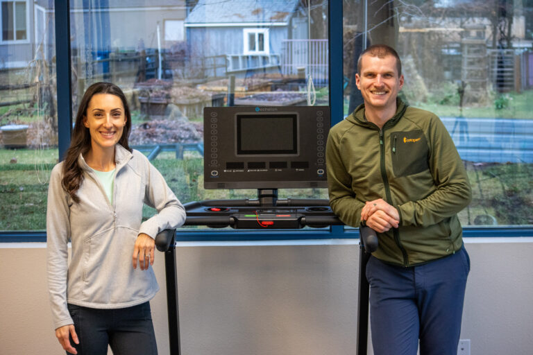 Physical therapists Ben Hoskyn and Maria Leibler smiling and leaning on opposite sides of a treadmill at Fit & Function Therapy Solutions.