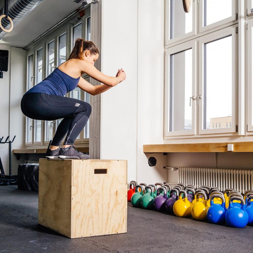 Woman performing a box jump during a strength training session with kettlebells in the background.